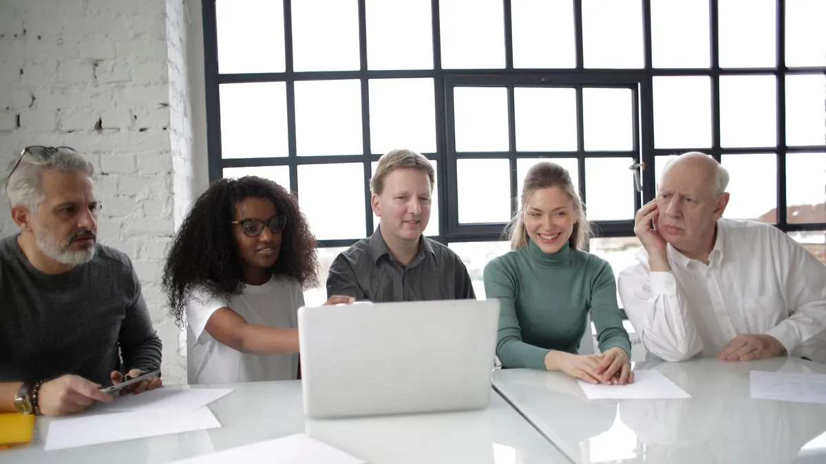 Group of multiracial coworkers of different ages discussing details of project while using computer at table in modern workplace