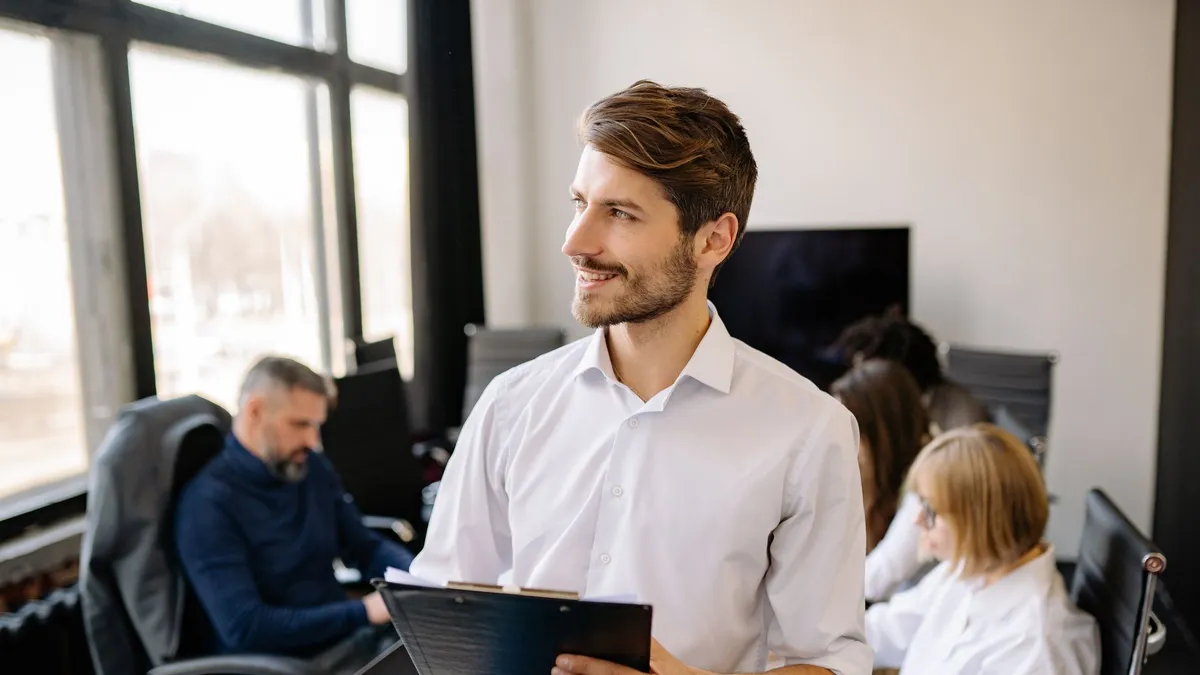 Businessman in a white dress shirt holding clipboard during a meeting in a modern conference room.
