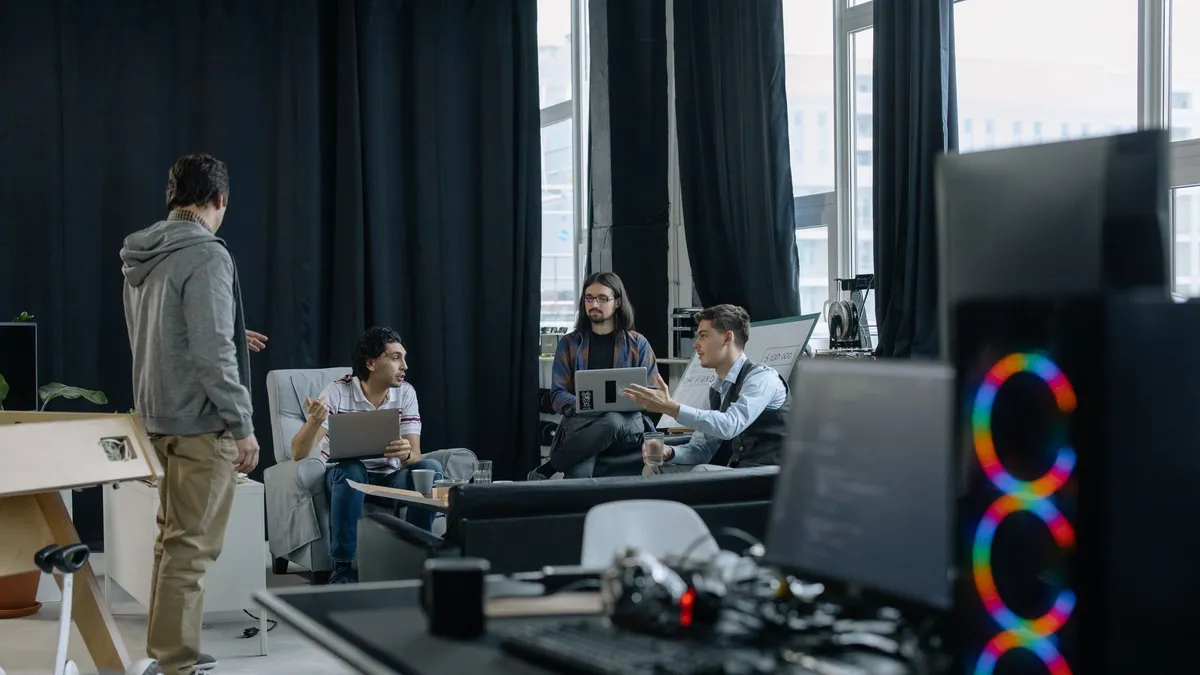 A group of professionals engaged in a casual meeting at a modern office, discussing projects on laptops and tablets.