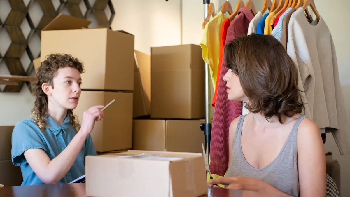 Two women engage in a business conversation surrounded by boxes and clothing in a fashion studio.