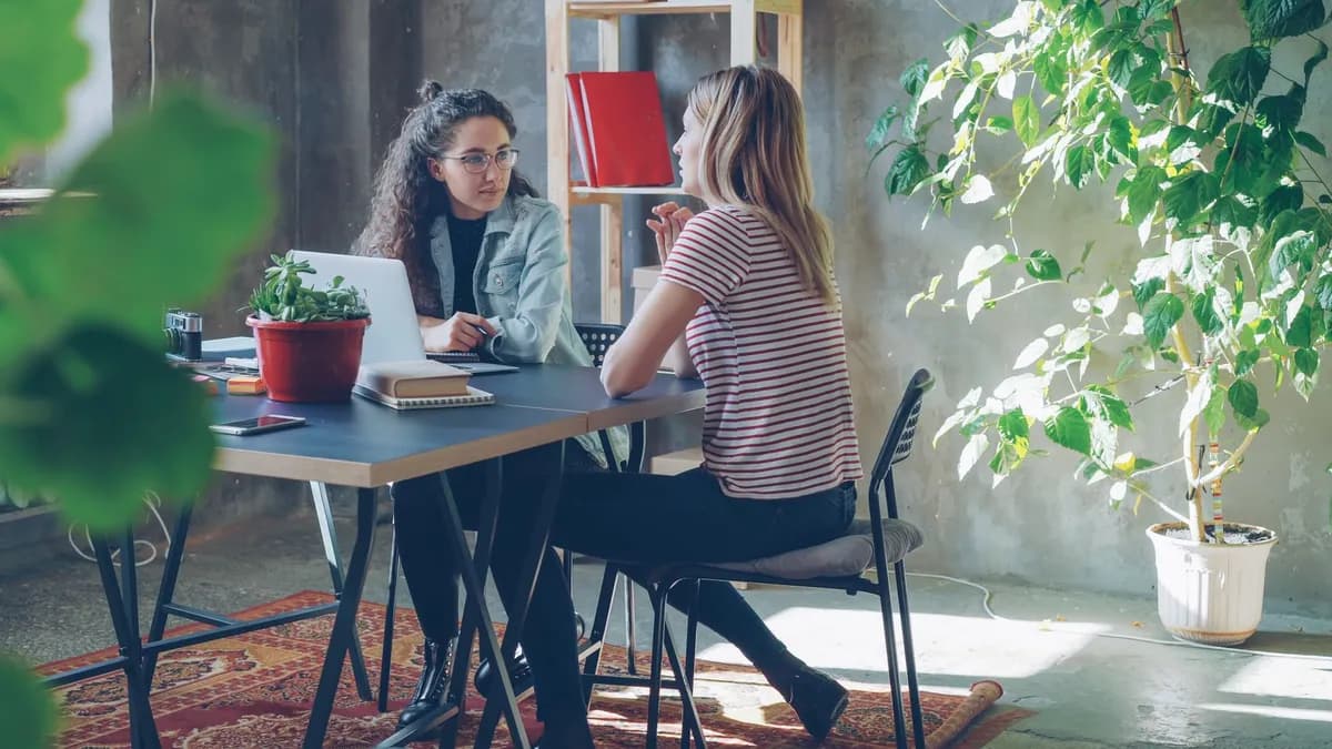 Two young women engaged in a discussion at a modern office with plants and sunlight.