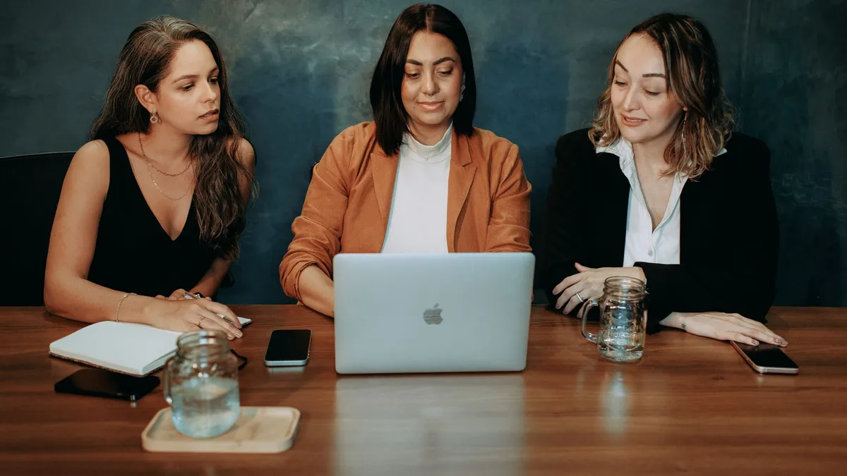 Three women engaged in a focused business meeting at a modern office setting with laptops and notebooks.