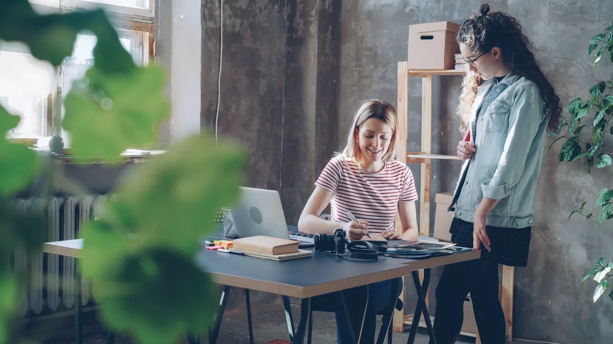 Two women working together at a desk in a modern, plant-filled office.