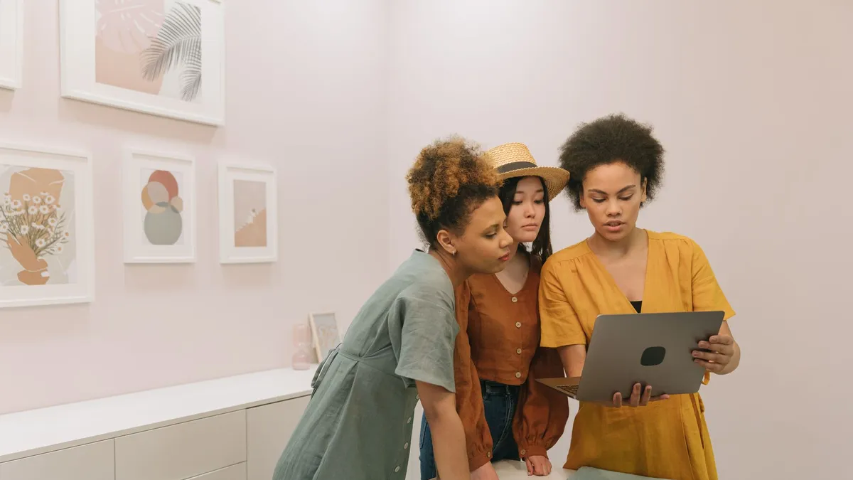 Three diverse women stand indoors, collaborating on a laptop surrounded by modern art.