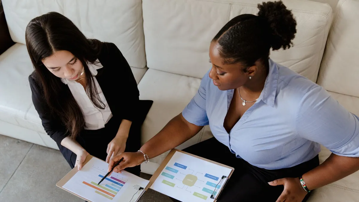 Two professional women discussing data charts during a business meeting on a couch.