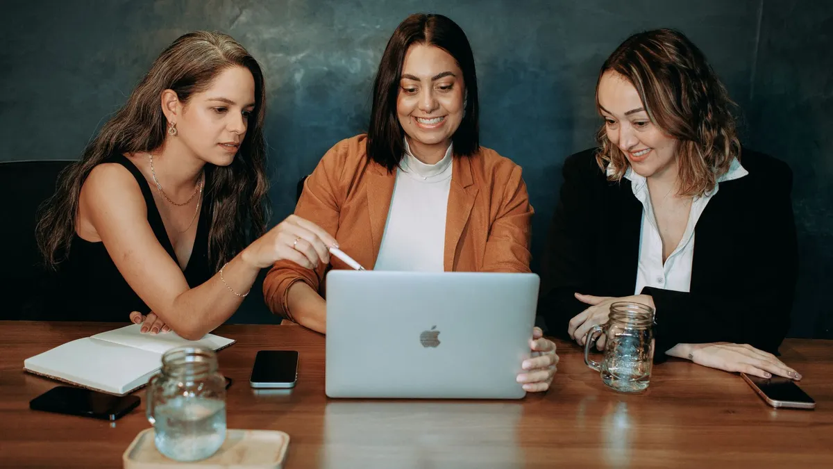 Three women in smart casual attire collaborate over a laptop in a business meeting.