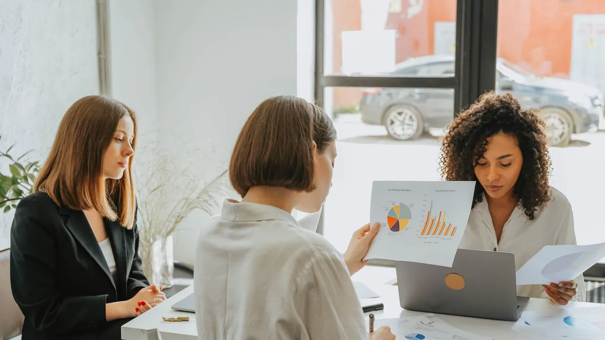 Three women engaged in a business meeting, discussing charts and data in a modern office setting.