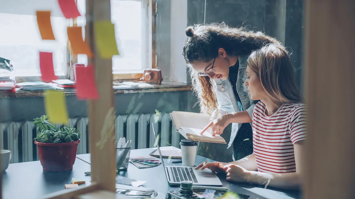 Two women collaborating in a cozy office space with laptops and notes.
