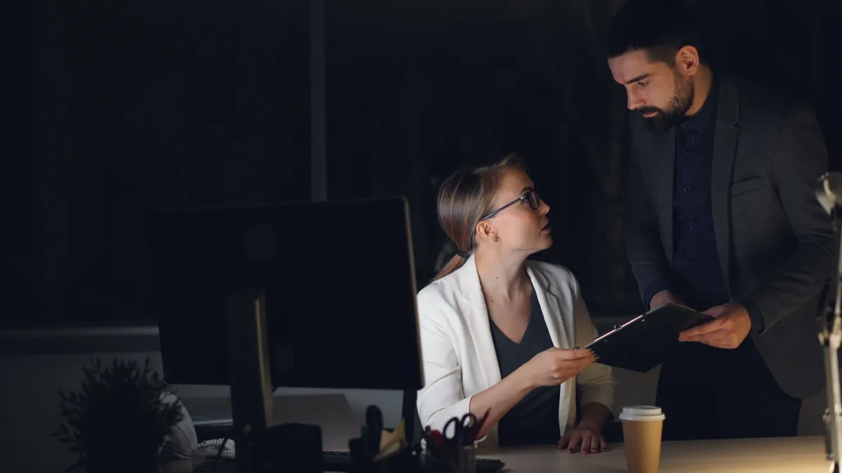 Two colleagues working late, discussing a project in a dimly lit office.