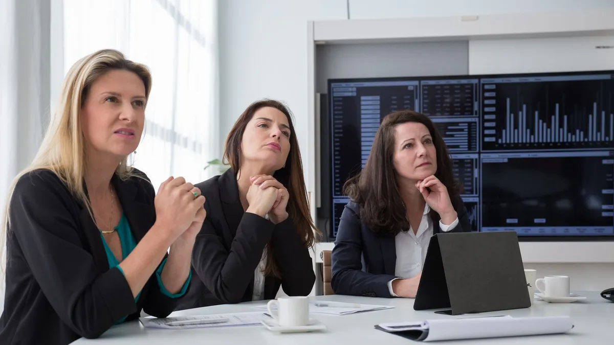 Three women in a business meeting room discussing charts and data on screens.