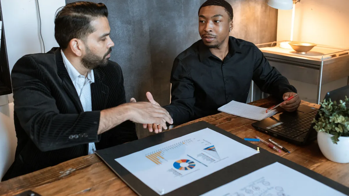 Two businessmen shaking hands over a desk with charts in a modern office.