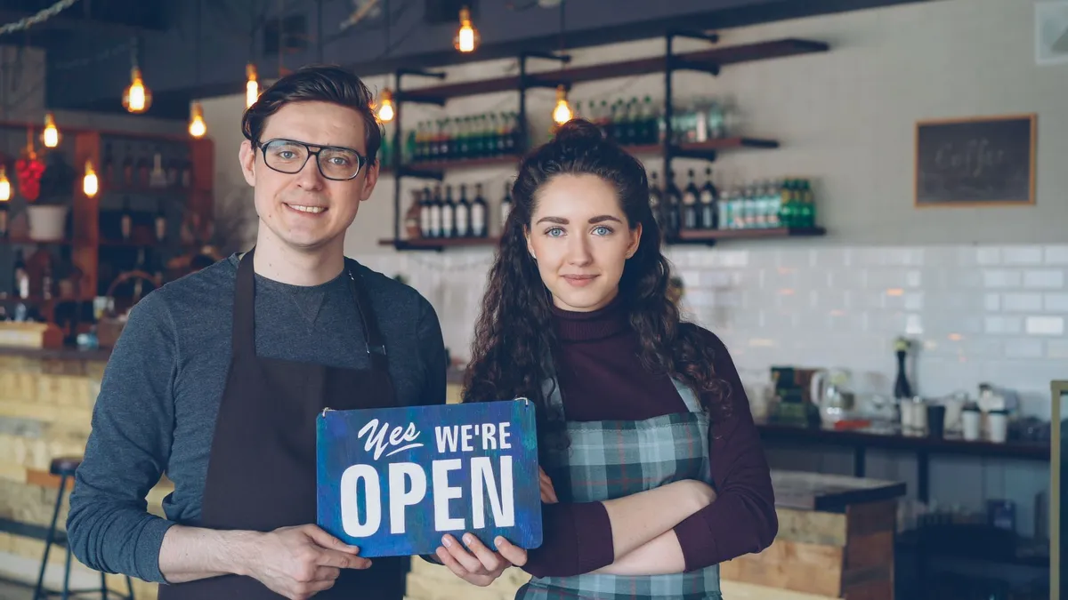 Friendly café owners holding an 'Open' sign welcoming customers into their modern coffee shop.