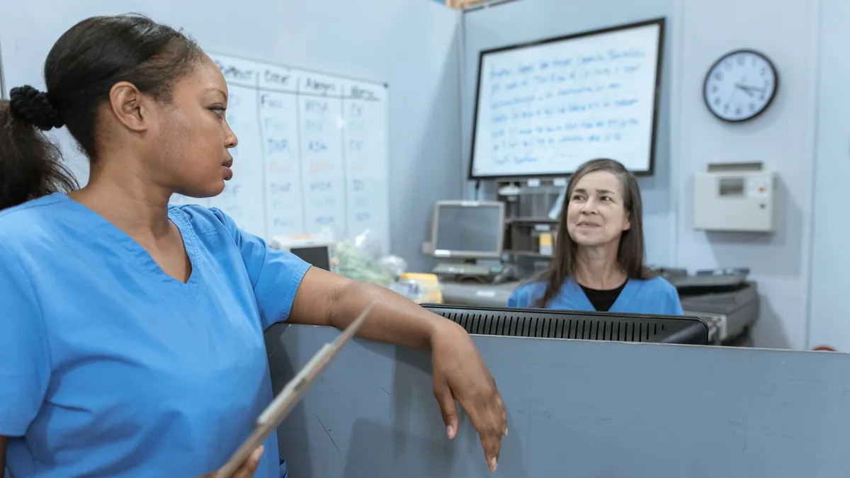 Nurses in scrub suits discussing medical tasks in a clinical environment.