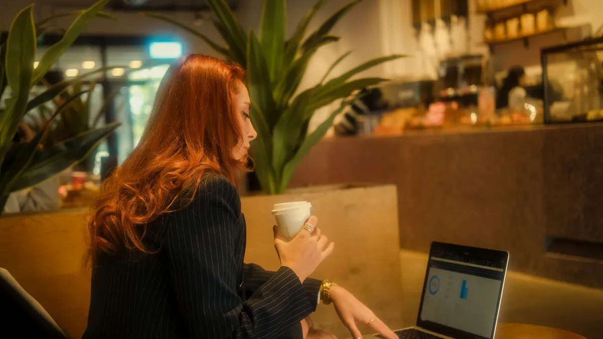 Redheaded businesswoman working on a laptop in a cozy café, holding a coffee.