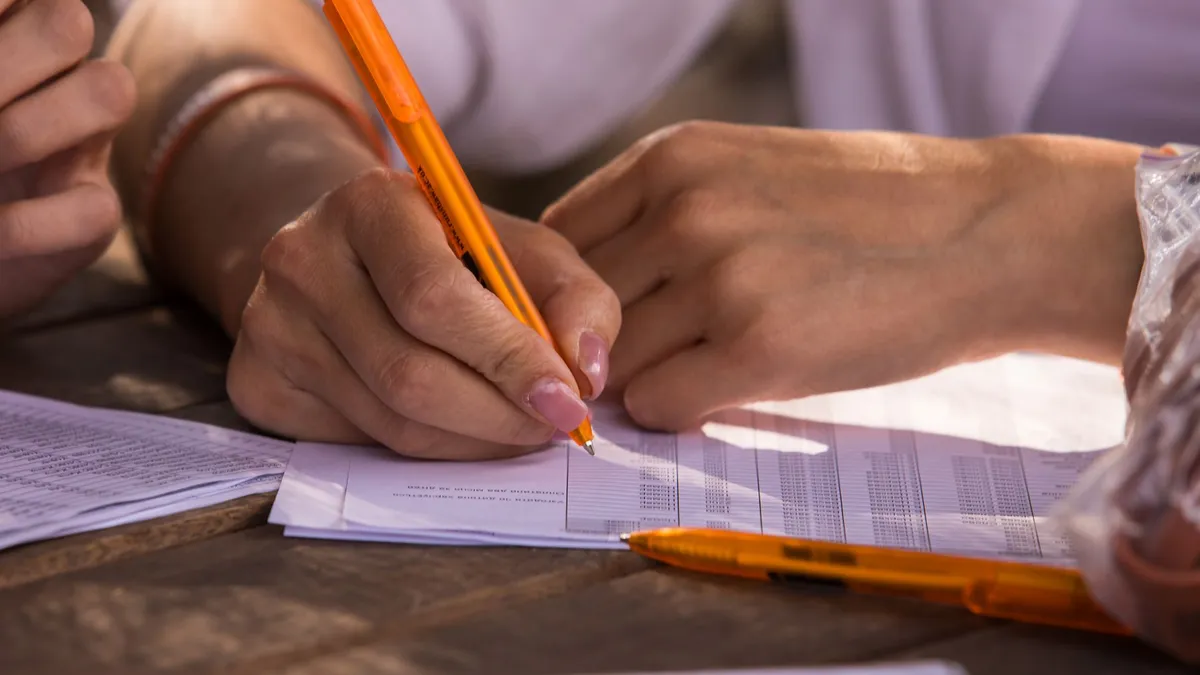 Close-up shot of hands writing on documents with orange pen, implying business or education setting.