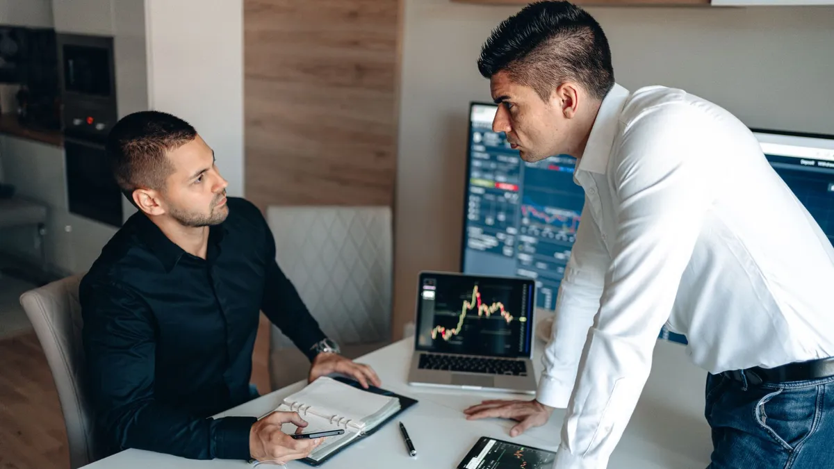 Two businessmen discuss stock market data on screens in a modern office.