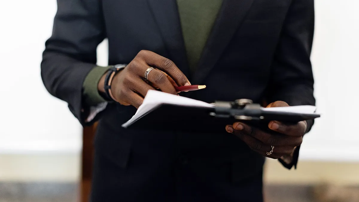 Close-up of an adult in a blazer holding a clipboard with documents and a pen indoors.