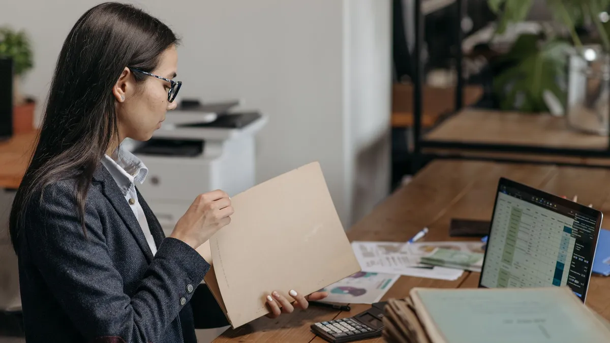 A professional woman reviewing documents at her office desk with a laptop and files.