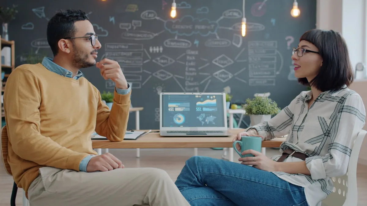 Two colleagues discuss ideas in a modern office with a chalkboard background.