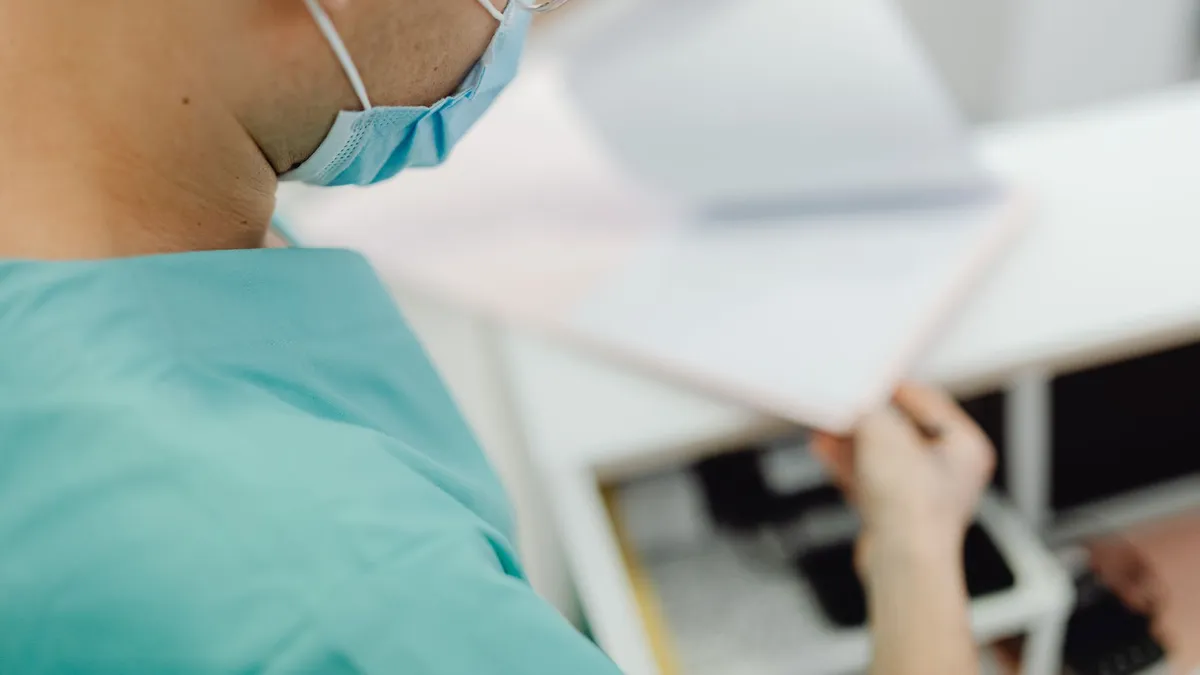 A medical professional in scrubs and mask examines documents in a clinical setting.
