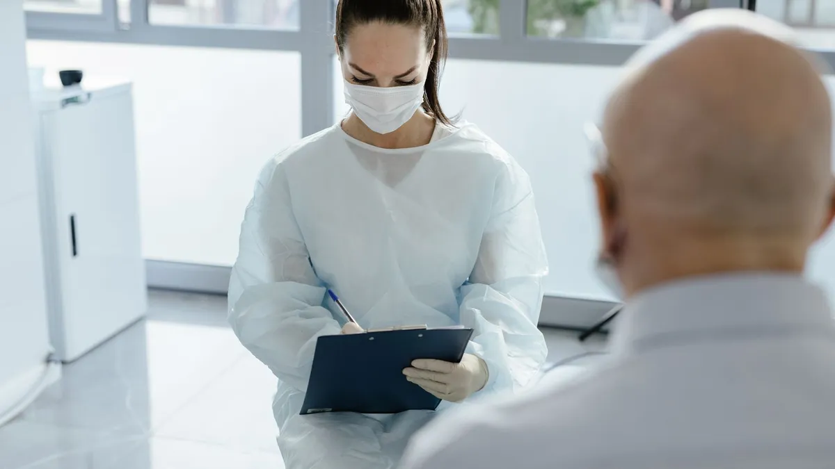 Doctor in protective gear taking notes during a medical consultation with a patient.