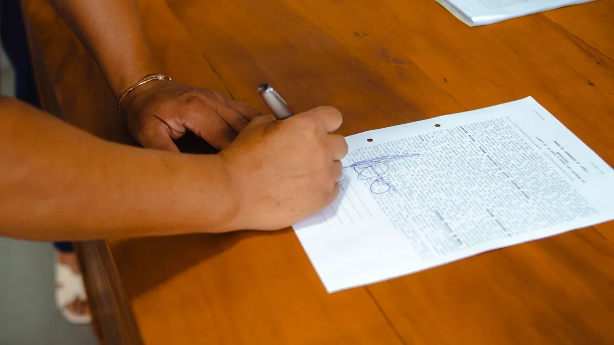 Close-up of hands signing a legal document on a wooden table indoors.