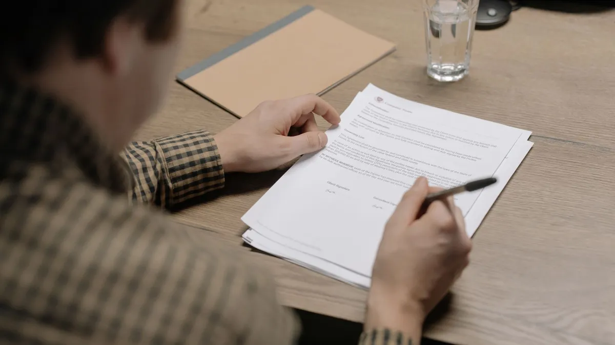 A businessman is reviewing a contract while holding a pen at a desk with a glass of water nearby.