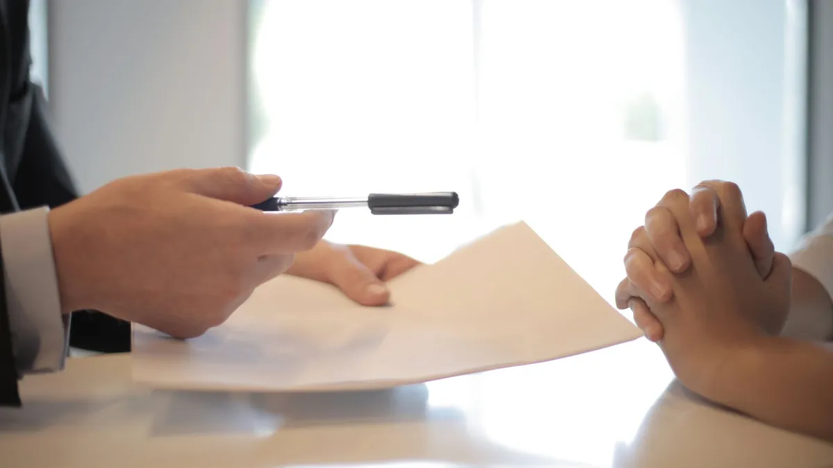 Side view of faceless formal man giving pen and paper to focused female with clenched hands at table on meeting