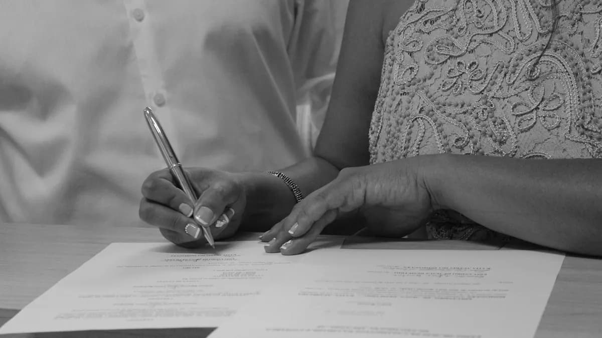 Black and white image of a couple signing documents, focusing on their hands and pen.