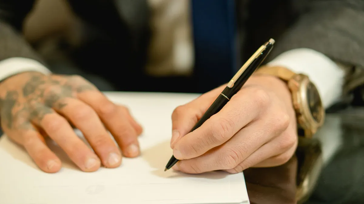 A close-up view of a businessman with tattoos signing a document at a desk.