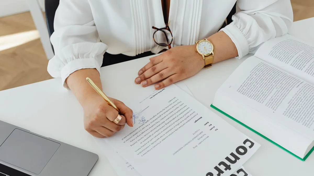 Close-up of a businesswoman signing a contract at her desk, with a laptop and book.