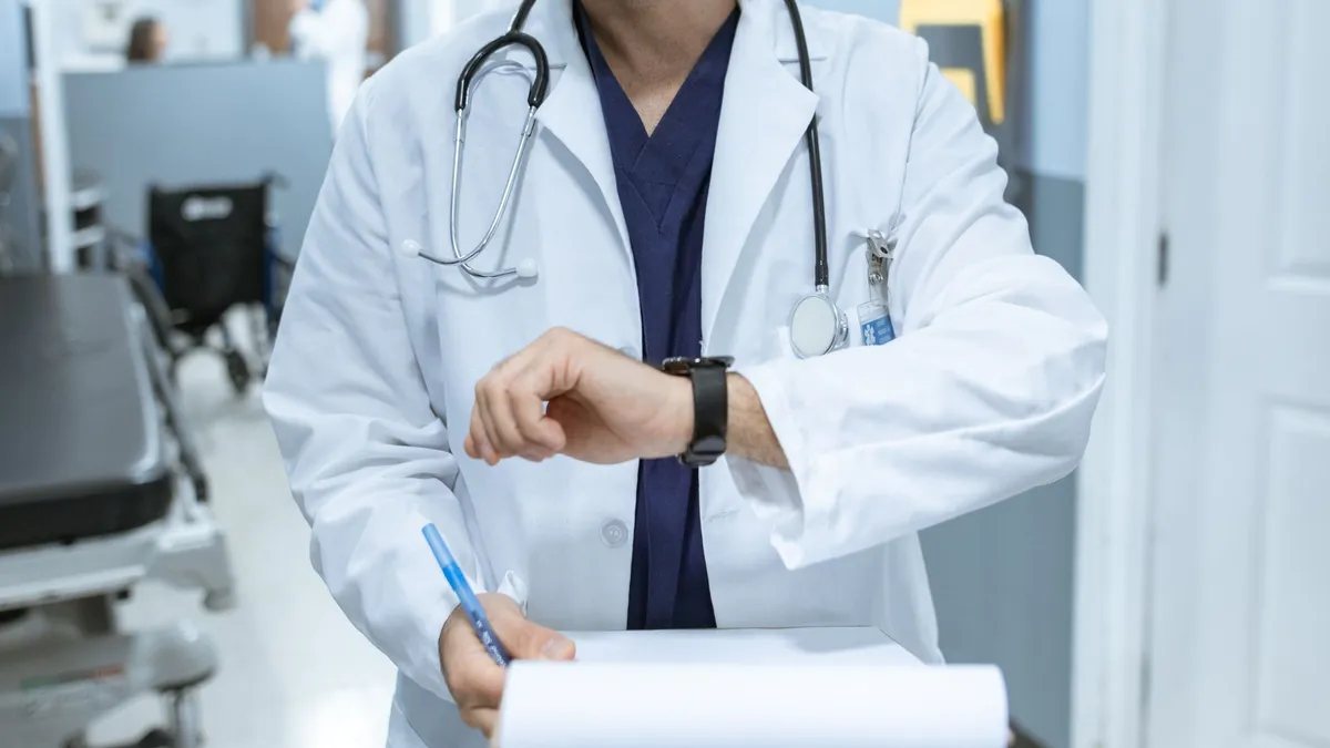 Doctor in lab coat checking wristwatch while holding medical chart in hospital corridor.