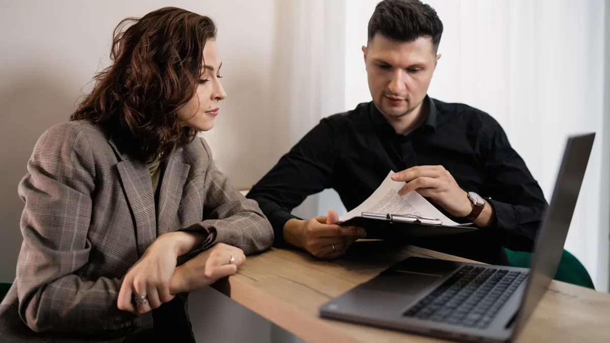Two professionals discussing documents at a desk with a laptop, focused on collaboration.