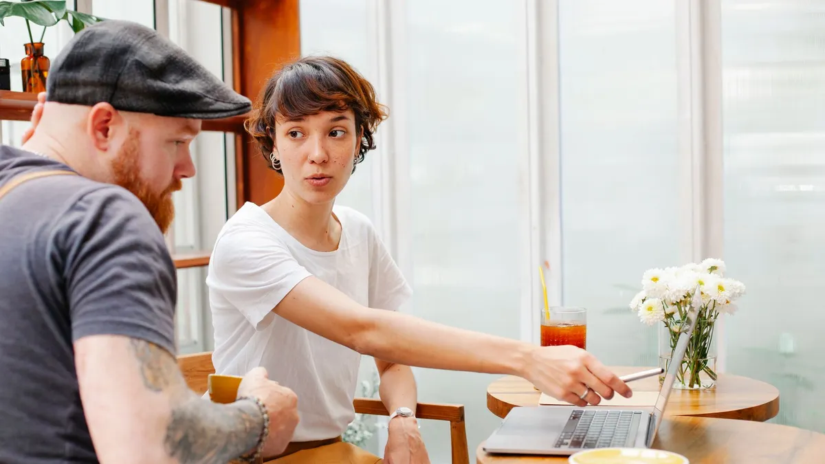 Young female barista pointing at netbook screen while talking to bearded male coworker in cafeteria