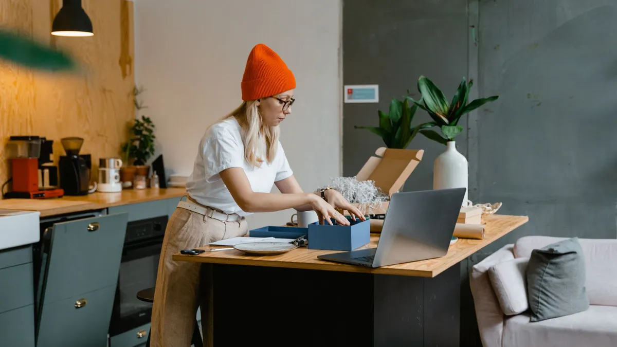 A young woman unpacks online shopping orders in a stylish modern kitchen, using her laptop for work.