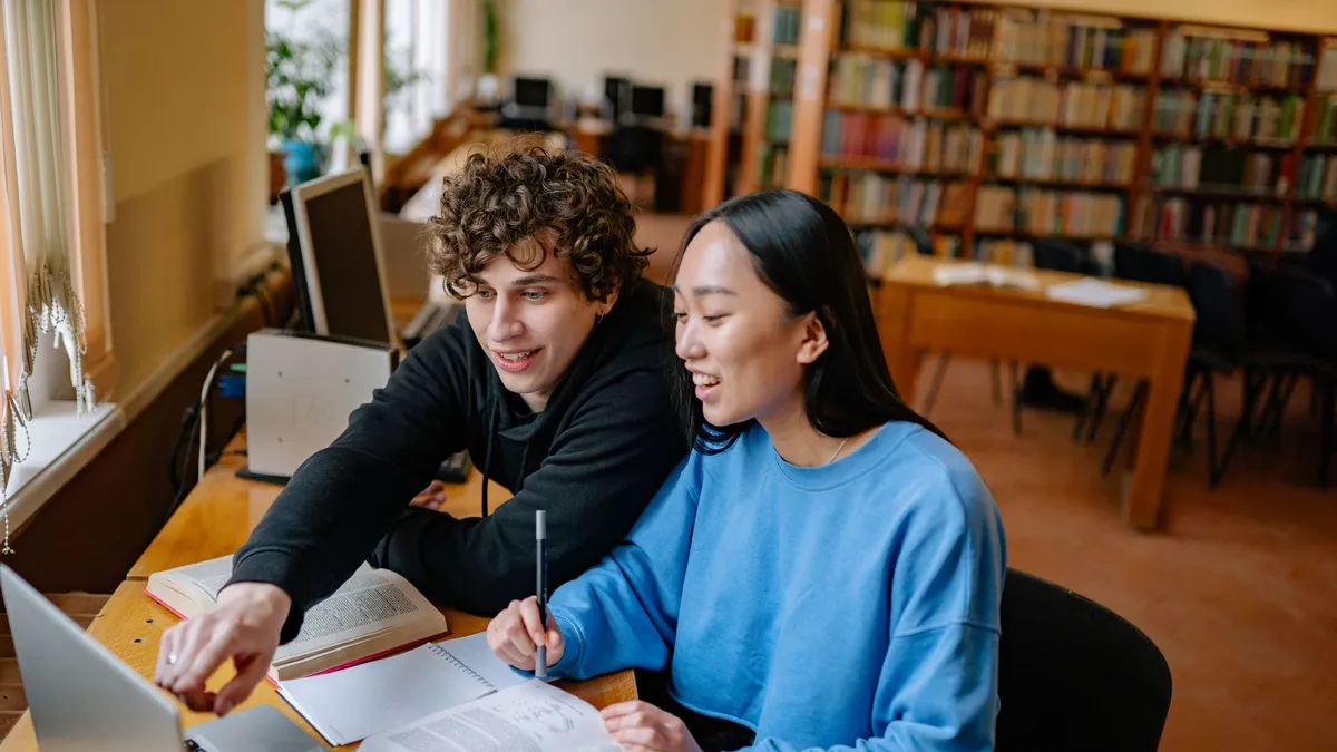 Two students studying together in a library with books and a laptop.