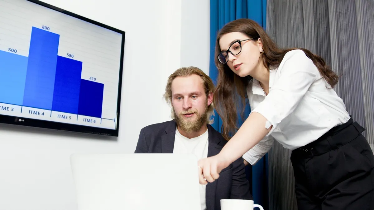 Two colleagues analyzing business data on a laptop with a presentation screen in the background.