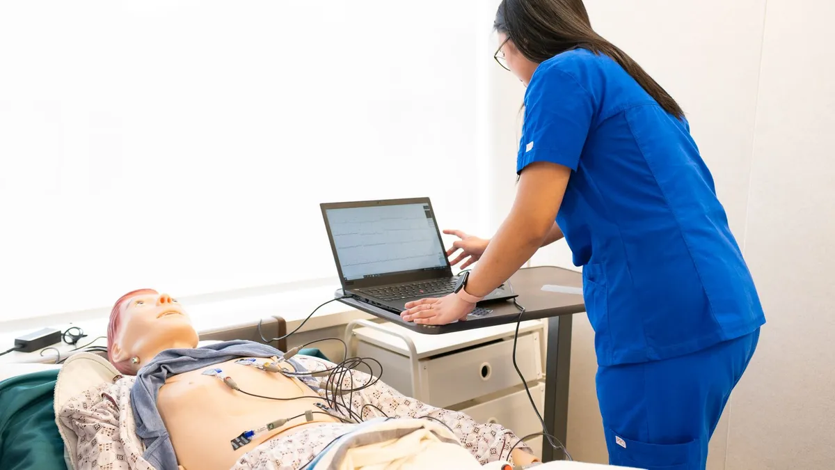 Nurse in training using a laptop with a medical dummy in a clinical simulation lab.