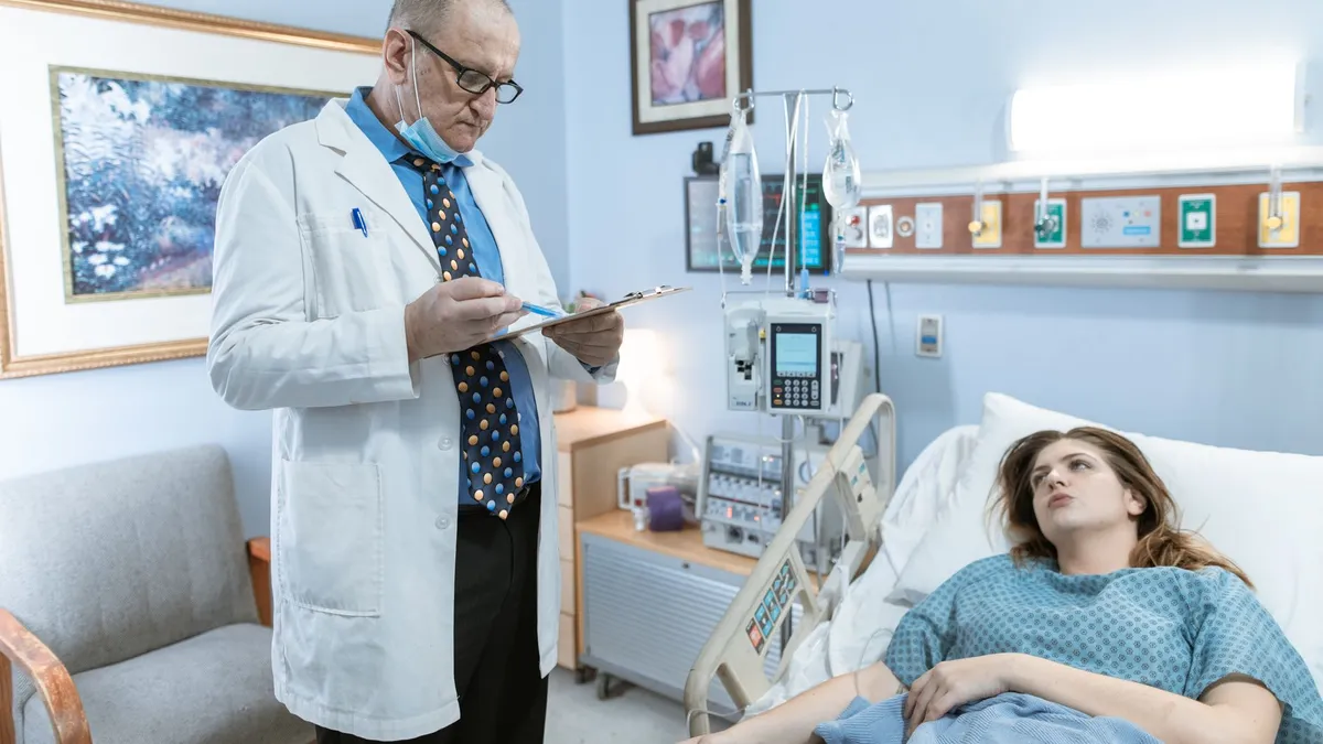 Doctor in white coat checking patient chart in hospital room with medical equipment.