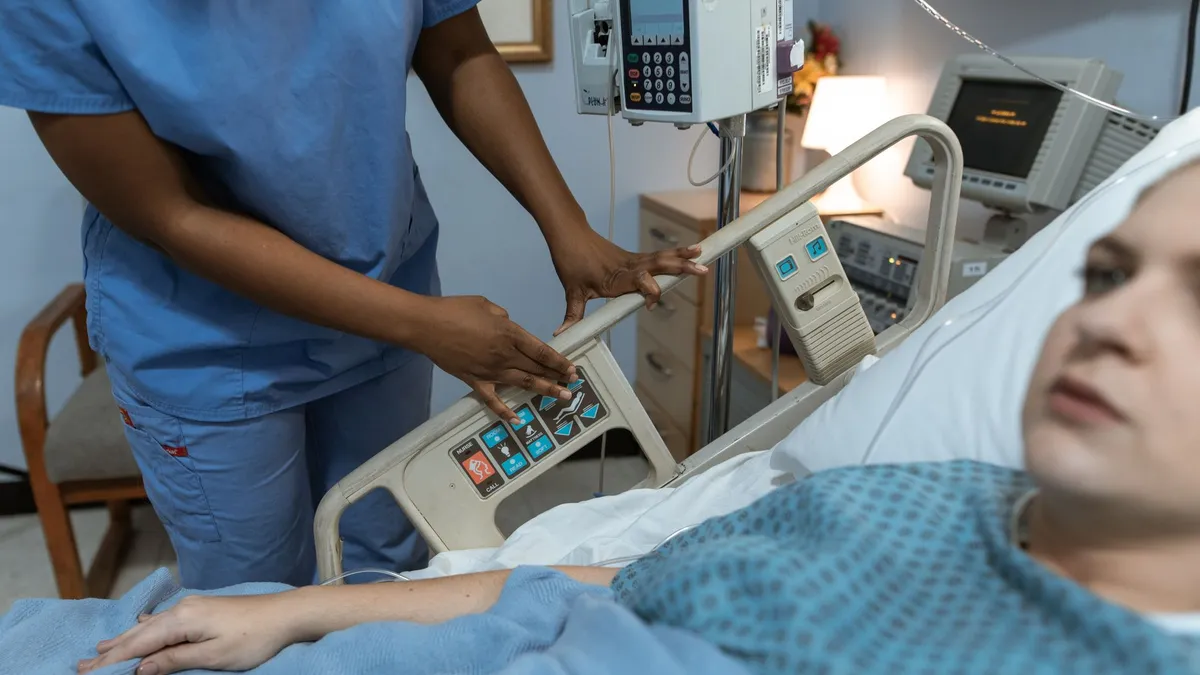 A nurse in scrubs assists a patient in a hospital bed with medical equipment nearby.