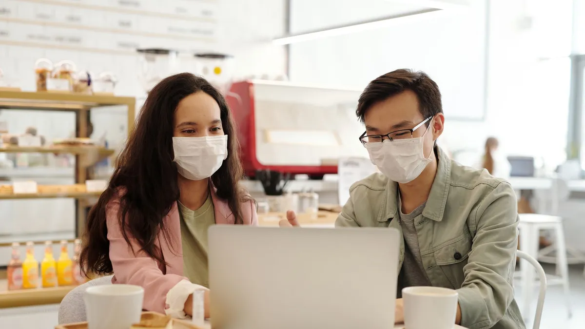 Two people wearing face masks discuss work on a laptop in a cafe setting.