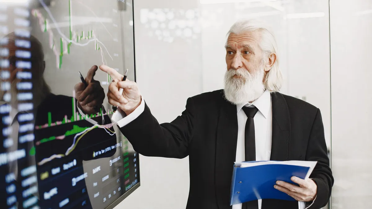 Elderly businessman pointing at stock market charts in an office setting, highlighting data analysis.