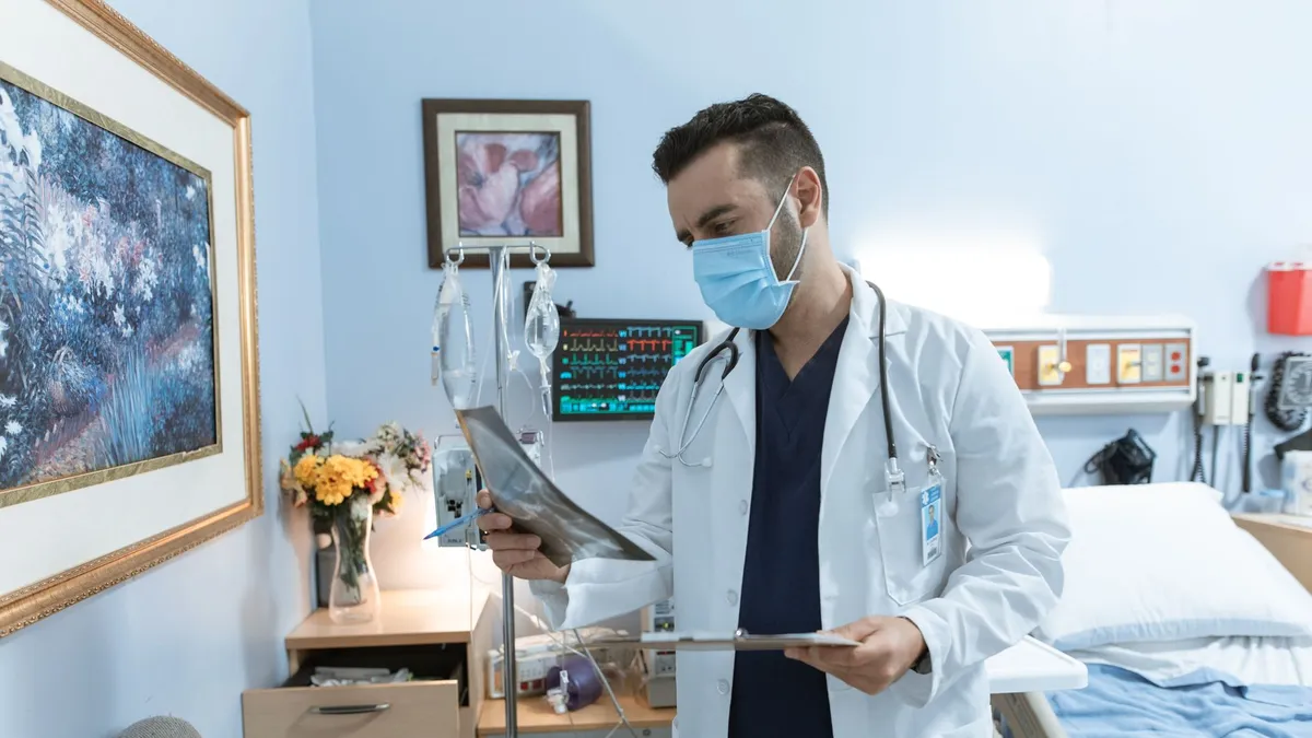 A male doctor wearing a face mask reviews an X-ray in a hospital room, focusing on patient diagnosis.
