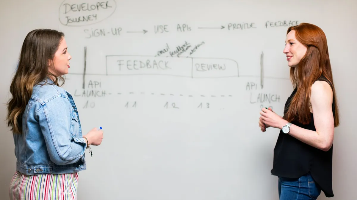 Two women engaging in a discussion about API development processes at a whiteboard.