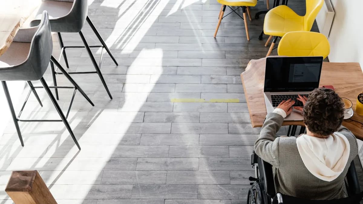From above of anonymous disabled female freelancer typing on netbook sitting at table while working remotely in modern spacious cafeteria