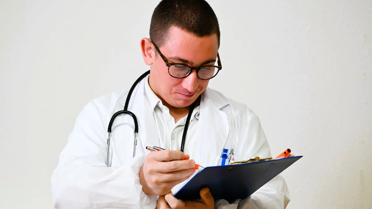 A male doctor in a white coat writes on a clipboard in a hospital setting, showcasing healthcare professionalism.