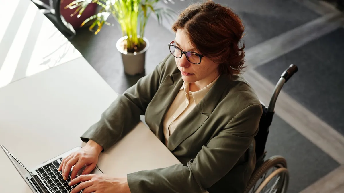 Professional woman in wheelchair working on laptop at a desk in an office setting, promoting inclusivity.
