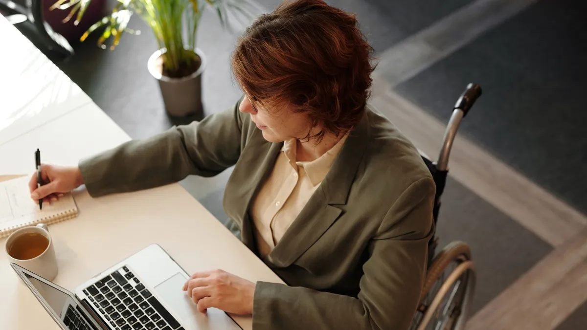 Top view of a businesswoman in a wheelchair working at her desk with a laptop and notebook.
