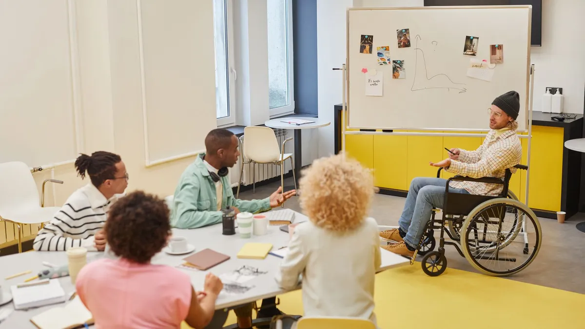 Diverse team engaged in a collaborative office meeting with a presenter in a wheelchair explaining ideas.