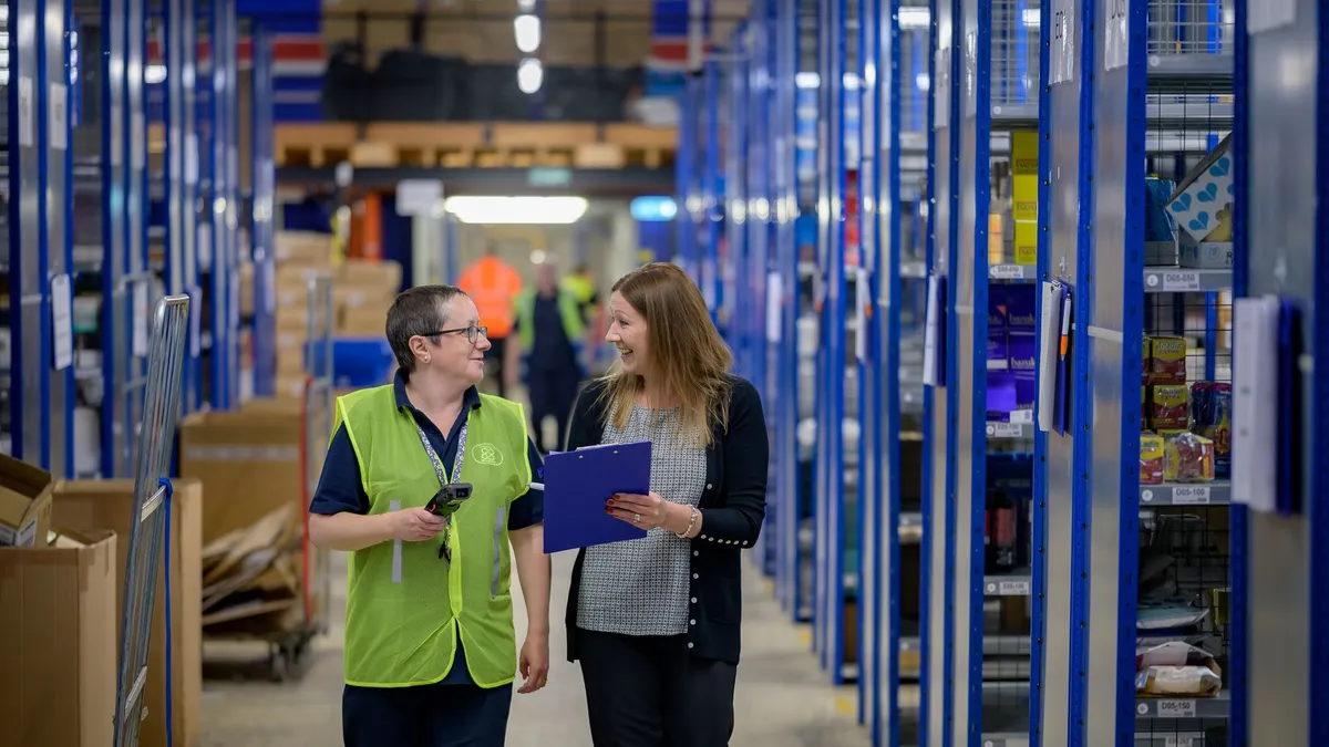 Two workers in reflective vests collaborate in a busy warehouse aisle.
